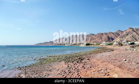 Paysage côtier au nord de Dahab, Sinaï, Égypte, regardant vers le sud - rivage ensoleillé avec des bâtiments lointains et la chaîne de montagnes escarpées s'élevant au-delà de th Banque D'Images