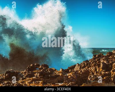 Violente vague océanique explose de façon spectaculaire au-dessus de roches déchiquetées, brun-rougeâtre, sous un ciel bleu clair Banque D'Images