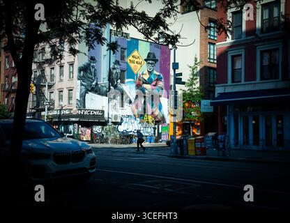 Tôt dimanche matin sur l'avenue A et East 12th Street dans l'East Village de New York. Un homme avec un café traverse East 12th Street. Une grande murale de l'artiste de rue brésilien Eduardo Kobra représentant Run-DMC, un groupe hip-hop originaire du Queens, New York, remplit le côté d'un bâtiment de quatre étages en arrière-plan. Banque D'Images