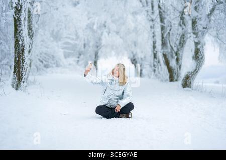 Femme assise sur un chemin enneigé capture selfie au milieu d'une forêt enneigée. Vêtue chaudement d'un manteau d'hiver, elle respire la joie et la sérénité dans le Banque D'Images