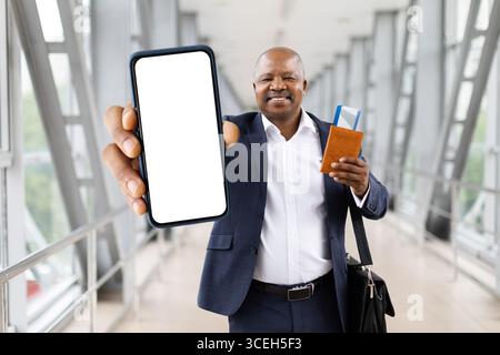 Homme d'affaires noir montrant smartphone avec écran vierge et tenant passeport à l'aéroport Banque D'Images