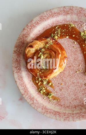 Une vue de haut en bas d'un rouleau de pâte feuilletée gastronomique, arrosé d'un glaçage au caramel sucré et saupoudré de pistaches hachées sur une assiette rose rustique. Banque D'Images