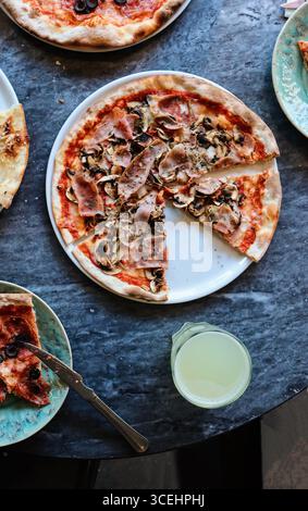 Pizza italienne fraîchement cuite garnie de jambon et de champignons, servie sur une assiette blanche sur une table rustique en pierre. Entouré d'autres tranches de pizza, olives Banque D'Images