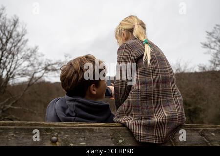 Garçon montrant quelque chose de drôle sur smartphone à la fille tout en étant assis sur une clôture en bois dans la forêt d'hiver Banque D'Images