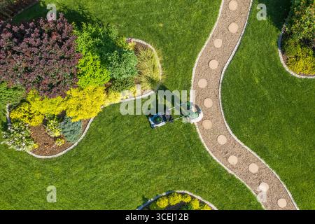 Une personne tond une pelouse verte luxuriante entourée de parterres de fleurs colorées dans un jardin bien entretenu. La scène capture une journée d'été avec un ciel dégagé. Banque D'Images