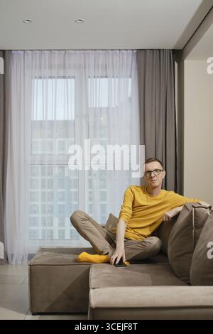 Un homme portant une chemise jaune et des chaussettes et un pantalon beige est assis confortablement sur un canapé marron, détendu dans un appartement moderne bien éclairé avec de grandes fenêtres et Banque D'Images