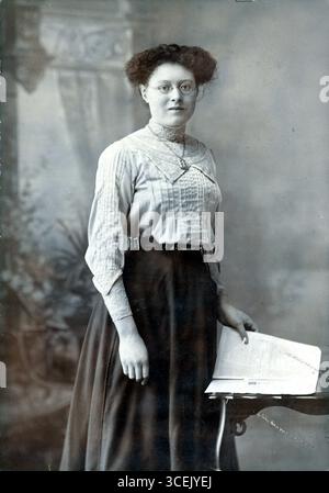 Une figure bespectacée se dresse devant un studio peint en toile de fond de colonnes et de draperies classiques, évoquant un raffinement doux. Vêtu d'une blouse rayée à col haut avec des accents de dentelle et d'une longue jupe sombre, le sujet repose une main sur une petite table portant un livre ouvert, peut-être un symbole d'apprentissage ou d'alphabétisation. La coiffure courte et bouclée et la posture confiante suggèrent la modernité du début du XXe siècle, mêlant présence intellectuelle et conventions formelles du portrait de l'époque. Extrait d'un lot de photographies originales d'albumen datant de la fin de l'époque victorienne et du début de l'époque édouardienne. Banque D'Images