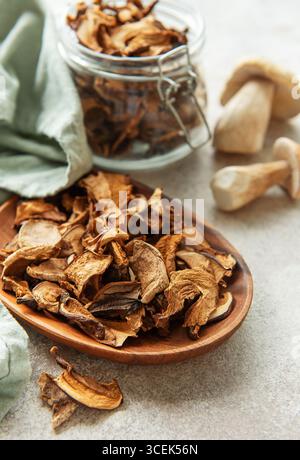Champignons porcini séchés se répandant d'une assiette en bois, avec des porcini frais et un pot de champignons séchés en arrière-plan Banque D'Images