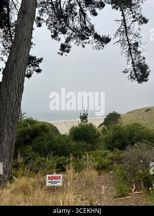 Un panneau d’avertissement rouge et blanc indiquant « danger – Hazardous Cliff » se dresse près d’une falaise abrupte surplombant la plage et l’océan Pacifique par une journée brumeuse Banque D'Images