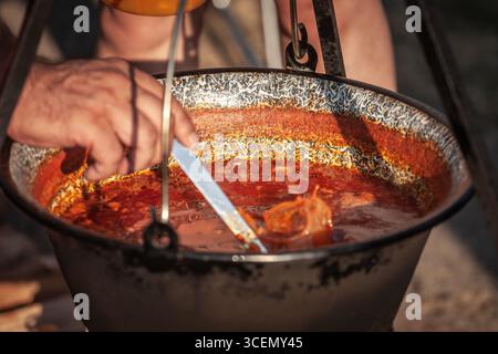 Une main remue la soupe de poisson riche en paprika dans un chaudron en émail accroché pour la cuisine en extérieur en Serbie. La scène montre riblja corba préparé dans un kotlic, avec Banque D'Images