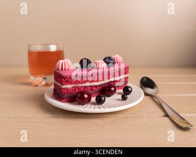 Une tranche de gâteau de velours rouge magnifiquement présentée avec glaçage rose et baies assorties sur le dessus, servie sur une assiette blanche. Le gâteau est accompagné d'un Banque D'Images