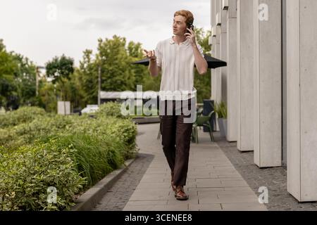 Ginger Man marchant le long de la rue en construisant et en parlant activement sur smartphone Banque D'Images
