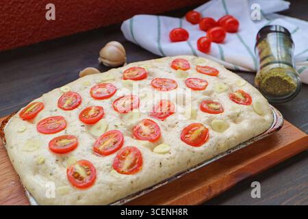 Des tranches d'ail et des tomates coupées ont été mises dans la pâte prête pour la cuisson du pain italien Focaccia Banque D'Images
