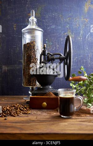 Tasse en verre de café noir fraîchement infusé sur une table en bois rustique avec du café moulu, des grains et un moulin vintage en arrière-plan, stylisé en confortable Banque D'Images
