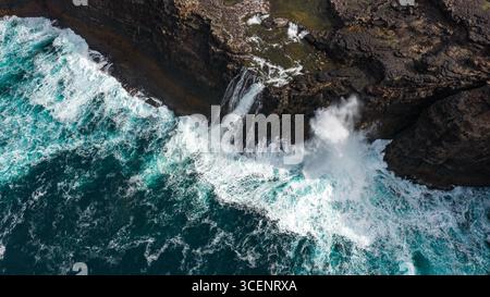 Vue aérienne d'un affrontement dramatique entre les falaises sombres de Trælanípa et l'océan turquoise, avec la cascade de Bøsdalafossur cascadant puissamment dans la mer, Miðvágur, Vágar, îles Féroé. Banque D'Images