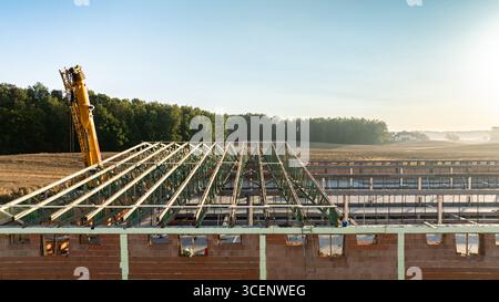 Vue tôt le matin d'un chantier de construction avec une structure de toit partiellement achevée faite de fermes d'acier vert et d'une grue jaune, entourée de fiel Banque D'Images