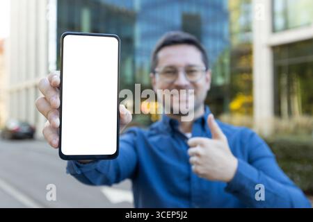 Homme souriant à l'extérieur tenant un écran de smartphone vierge vers l'appareil photo tout en donnant un pouce vers le haut, indiquant la satisfaction ou mettant en valeur l'espace pour le contenu personnalisable. Banque D'Images