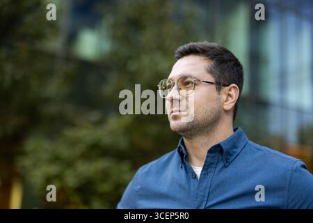 Un jeune professionnel confiant portant des lunettes debout à l'extérieur près d'un bâtiment moderne. Les environs comprennent des arbres et un extérieur en verre, créant une atmosphère professionnelle mais décontractée. Banque D'Images