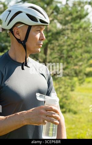 Cycliste debout avec une bouteille d'eau portant un t-shirt gris et se détendre à la forêt. Athlète prenant une pause. Journée d'activité extérieure. Saison estivale Banque D'Images