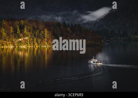 Vue d'un bateau glisse sur le lac tranquille reflétant le feuillage d'automne vibrant sur fond de montagnes enveloppées de brume, Hallstatt, Autriche. Banque D'Images