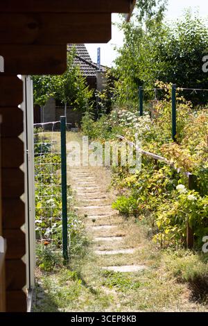 Un étroit chemin bordé de verdure et de fleurs mène à travers un jardin. Les structures en bois sont visibles en arrière-plan sous un ciel bleu clair. Banque D'Images