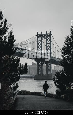 New York, États-Unis - 25 décembre 2023 : vue du pont de Manhattan alors qu'une figure solitaire marche vers l'East River, encadrée par les silhouettes sombres d'arbres. Banque D'Images
