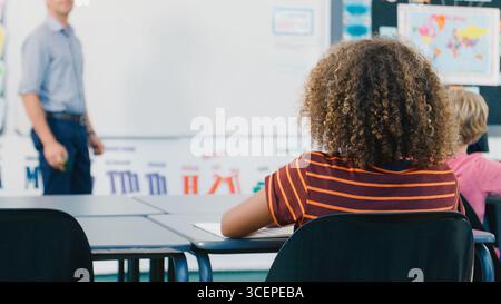 Vue arrière d'un élève de lycée assis au bureau pendant la leçon avec un professeur au tableau blanc Banque D'Images
