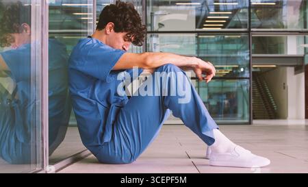Jeune médecin homme fatigué de porter des gommages corporels assis contre le mur dans la construction de l'hôpital moderne Banque D'Images
