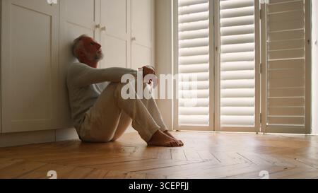 Homme âgé déprimé souffrant de mauvaise santé mentale assis sur le plancher à la maison Banque D'Images