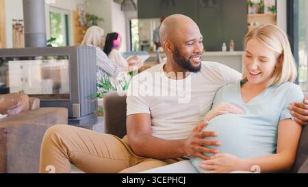 Couple multi-racial avec femme enceinte sur canapé à la maison avec famille multi-génération en arrière-plan Banque D'Images