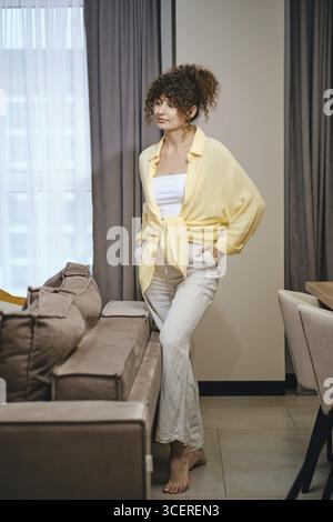Une femme est habillée de façon décontractée dans une chemise jaune et un pantalon beige tout en se tenant debout à côté d'un canapé dans un intérieur élégant. L'espace est lumineux, avec larg Banque D'Images