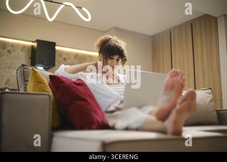 Une jeune femme est assise confortablement sur un canapé, les pieds vers le haut, en utilisant son ordinateur portable pour les appels vidéo en ligne dans un salon lumineux décoré de neutres chauds Banque D'Images