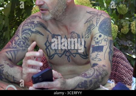Jeune homme sur téléphone portable et tatouages sur les bras et le haut du corps, assis dans le jardin, Bade-Wuerttemberg, Allemagne Banque D'Images