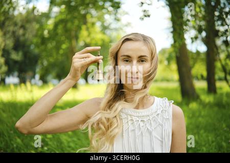 Une jeune femme se tient dans un parc vert vibrant, souriant en tenant une petite cloche. La lumière du soleil filtre à travers les arbres, éclairant son h qui coule Banque D'Images