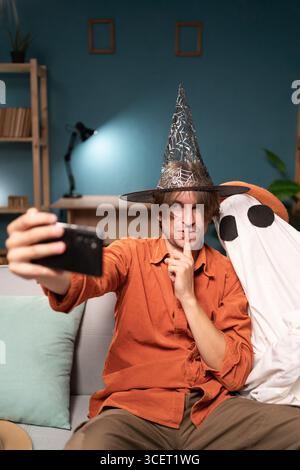 Joyeux homme de sorcière pointant shh geste et fantôme effrayant dans le costume d'halloween faisant selfie sur smartphone assis sur le canapé dans le salon. Halloween Banque D'Images