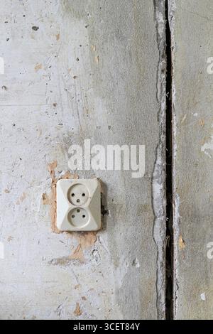 Une photo en gros plan d'une vieille prise électrique sur un mur de béton rugueux avec des fissures, du plâtre écaillé et des dommages visibles. La douille usée et non finie Banque D'Images