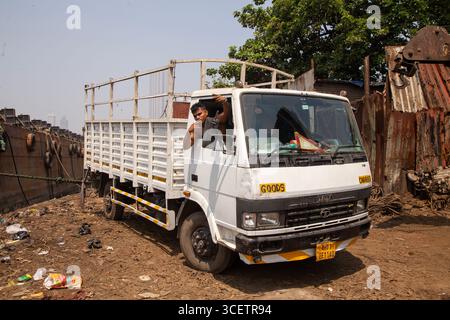 Un camion de transport de marchandises blanches est garé sur un terrain industriel poussiéreux, entouré d'objets jetés et de hangars métalliques, sous la lumière du jour avec des arbres Banque D'Images
