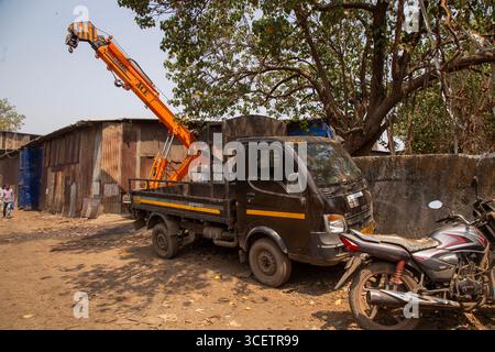 Un camion utilitaire équipé d'un bras de grue orange étendu est stationné à côté des motos dans un espace extérieur poussiéreux près d'un ancien hangar et d'un grand arbre Banque D'Images