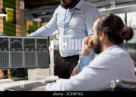 Gestionnaire supervisant le travail de l'analyste de risque utilisant la plate-forme de négociation pour examiner les cours des actions boursières. Le chef d'équipe et l'employé de la société de soutien analysent les rapports de portefeuille, vérifiant les risques financiers Banque D'Images