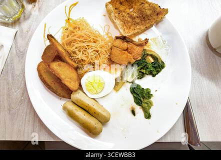 Vue de dessus assiette d'aliments de petit déjeuner assortis, y compris les œufs, nouilles, quartiers de pommes de terre, légumes verts, saucisses, et du pain sur une table en bois Malacca malaisie Banque D'Images