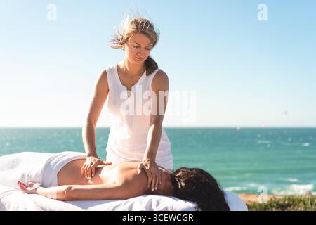Masseur professionnel redonnant massage à la femme couchée sur la table près de la mer contre le ciel bleu. Soin spa pendant les vacances d'été. Relaxation, mode de vie sain et soins corporels Banque D'Images