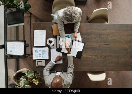 Femme caucasienne d'âge moyen et homme noir d'âge moyen assis à une table en bois serrant la main sur des documents d'affaires et du café, vue de dessus montrant la réunion d'accord professionnelle Banque D'Images
