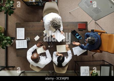 Groupe de médecins multiethniques d'âge moyen et senior et un homme caucasien d'âge moyen assis autour de la table discutant des documents médicaux et utilisant un ordinateur portable pendant la réunion, vue de dessus Banque D'Images