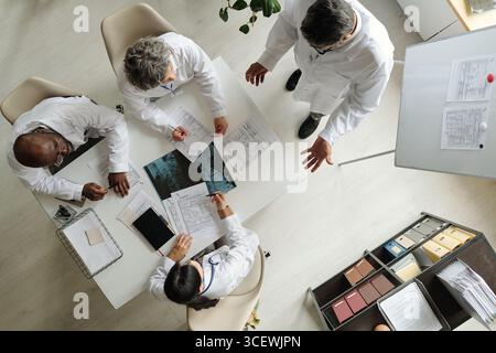 Groupe de médecins multiethniques d'âge moyen discutant des documents médicaux et analysant des images de rayons X autour de la table dans le bureau, debout et assis, collaborant sur le diagnostic du patient Banque D'Images