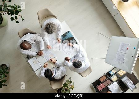 Groupe de médecins multiethniques d'âge moyen discutant des documents médicaux autour de la table, collaborant sur les soins aux patients, examinant des graphiques et prenant des notes, vue de dessus des professionnels de la santé Banque D'Images