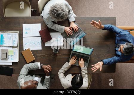 Groupe de professionnels multiethniques d'âge moyen collaborant autour d'une table, discutant des graphiques financiers et utilisant des ordinateurs portables, vue de dessus montrant le travail d'équipe et l'engagement actif Banque D'Images