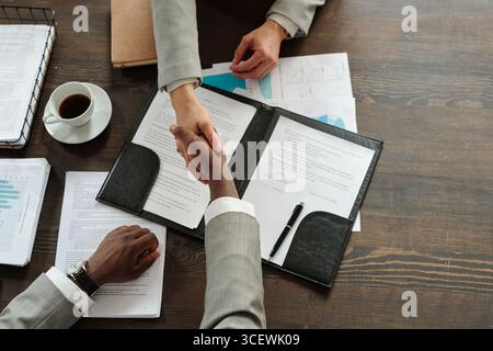 Femme caucasienne et homme noir serrant la main sur les documents contractuels sur le bureau en bois, les deux portant des vêtements d'affaires, tasse à café et graphiques financiers visibles, composition de vue de dessus Banque D'Images