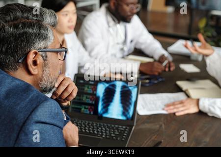 Homme caucasien d'âge moyen examinant l'image de radiographie thoracique sur ordinateur portable avec une femme asiatique et un homme noir dans des uniformes médicaux discutant le diagnostic à table pendant la réunion, les mains faisant des gestes dans la conversation Banque D'Images