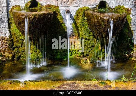 Trois becs en pierre avec de l'eau claire coulent d'une fontaine de village rustique couverte de mousse et de lumière du soleil. Banque D'Images