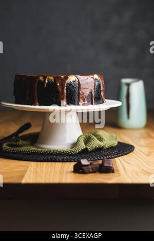 Elégant cheesecake avec une croûte de biscuits foncée, surmonté d'une riche ganache au chocolat, coiffé sur un support à gâteaux blanc avec un décor rustique. Banque D'Images